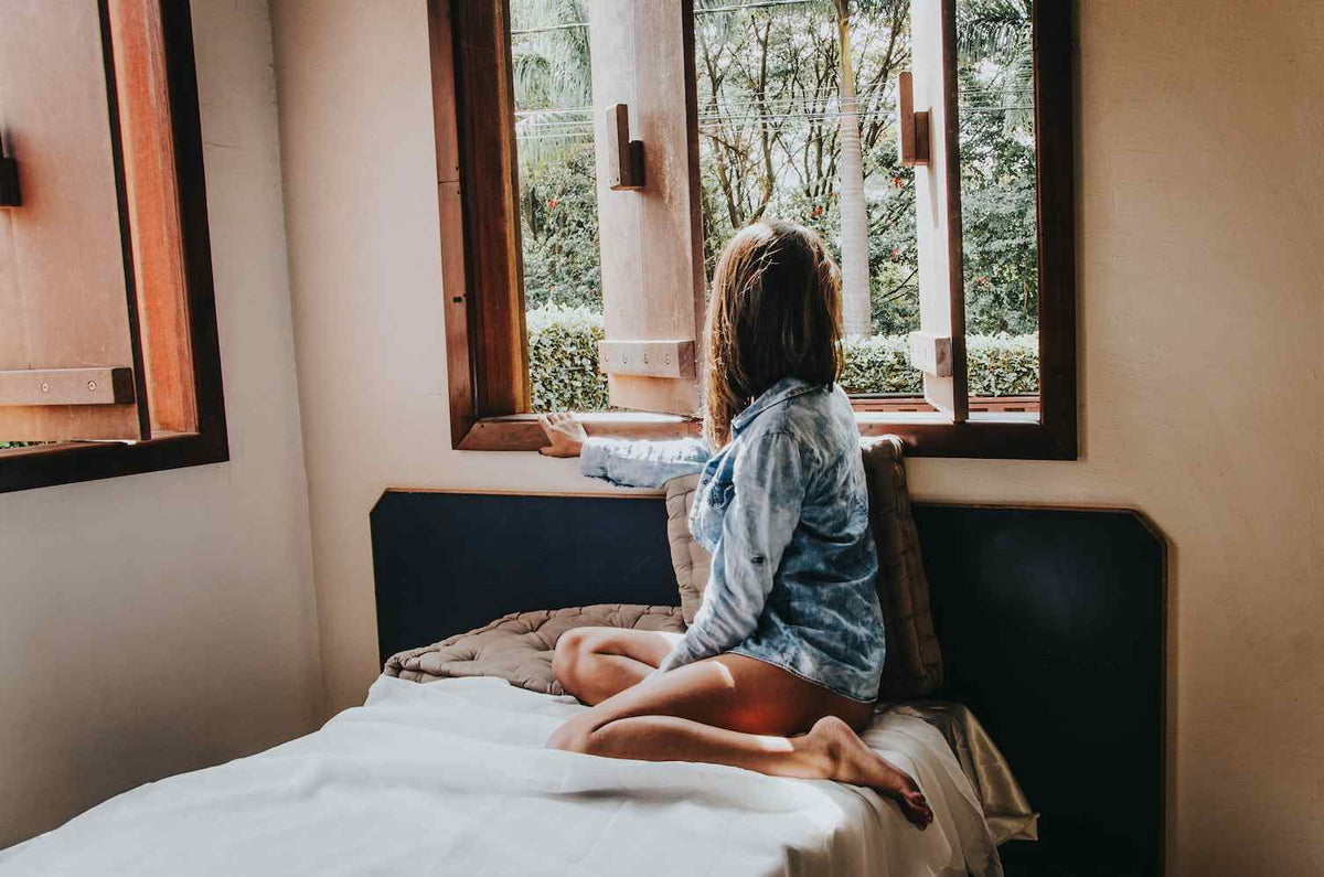 Girl wearing a denim shirt sitting on a bed looking out at the woods
