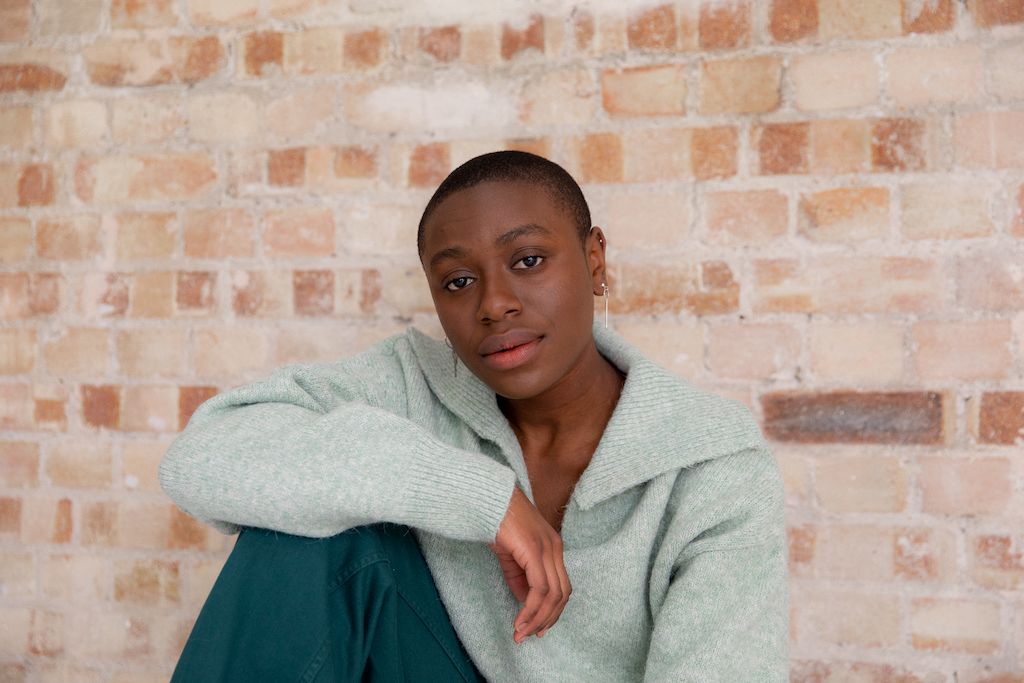Non Binary person sitting wearing a teal wool jumper and dark blue trousers